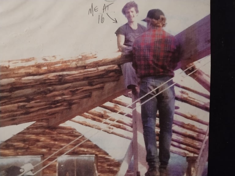 Two people standing on wooden log cabin structure during construction, vintage photograph dated 1978