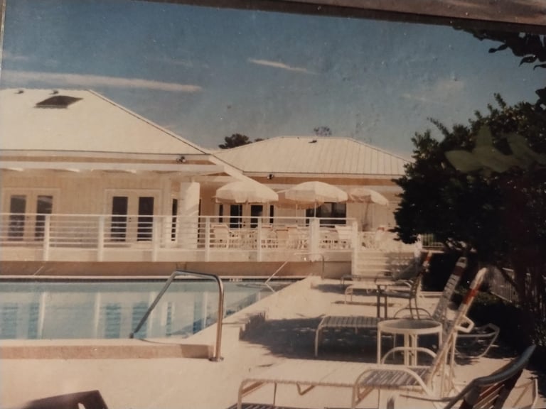 Outdoor pool facility with white building, lounge chairs, and landscaping on sunny day