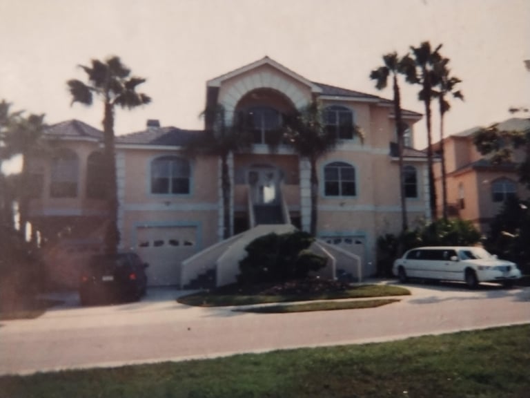 Two-story beige house with palm trees, white vehicle in driveway, and garage doors visible from street