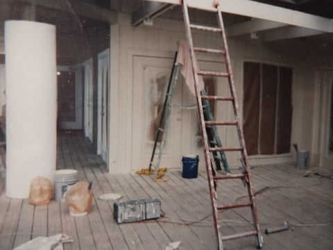 Interior construction site with wooden flooring, extension ladder, paint cans, and doorways in an unfinished room