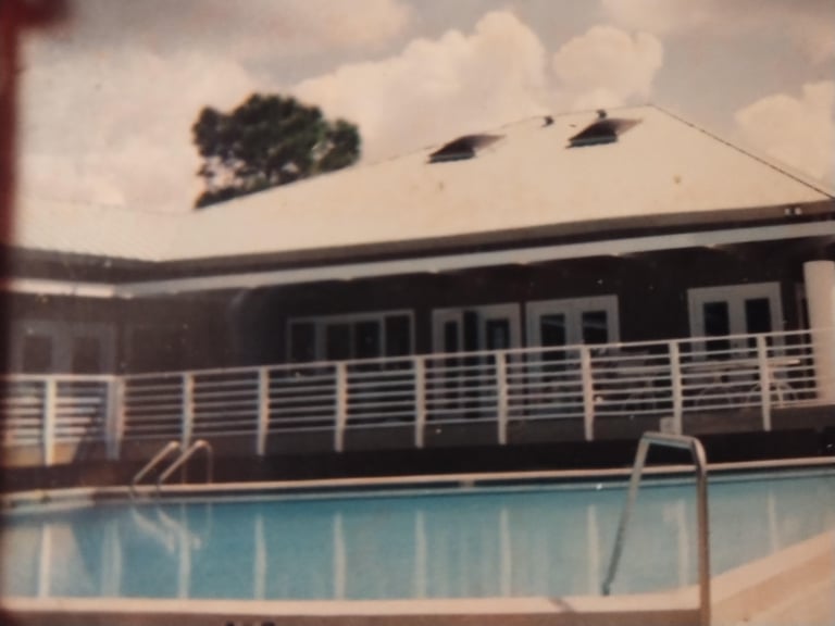 Vintage poolside view of a mid-century building with white railings overlooking a blue swimming pool under cloudy sky