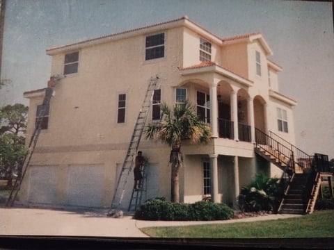 Beige two-story coastal home with elevated foundation, double garage, palm trees, and wooden deck stairs