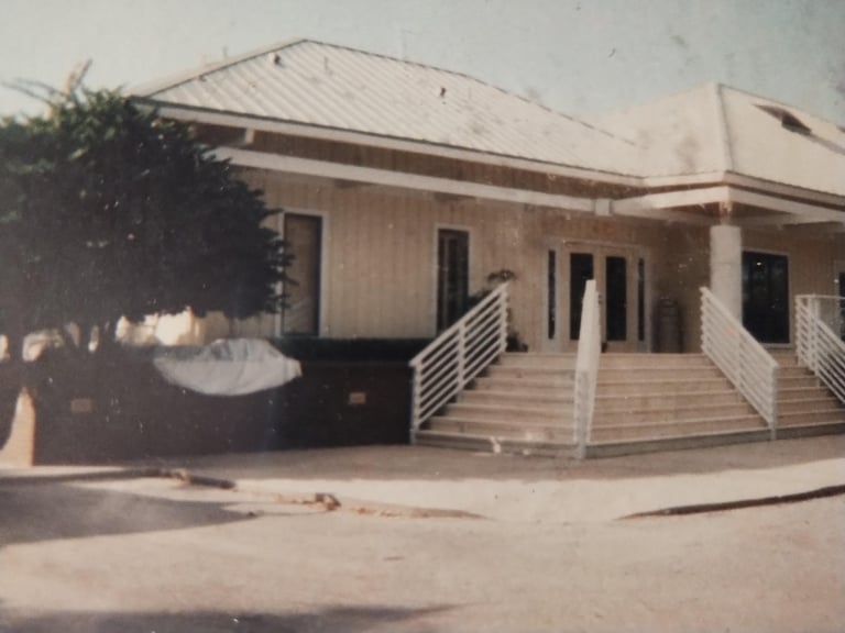 Exterior view of a single-story tan building with metal roof, white railings, and wooden ramp entrance, vintage photograph